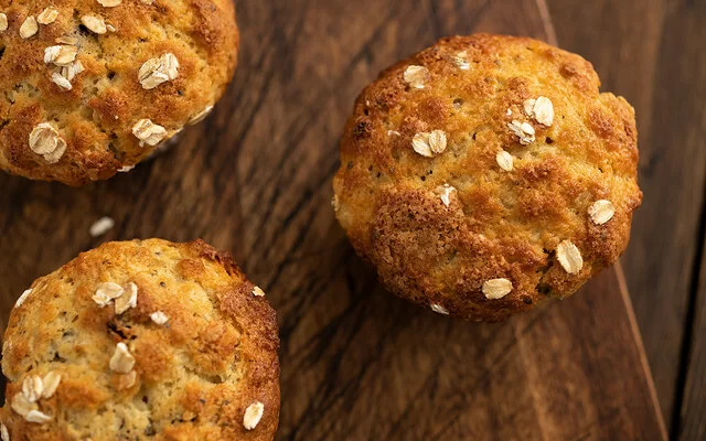 Aus der Vogelperspektive sind drei frische Muffins mit Haferflocken zu sehen. Sie stehen auf einem rustikalen Holzbrett.
