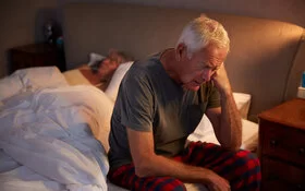 Ein älterer Mann sitzt mit leicht gesenktem Kopf auf einem Bett und stützt den linken Arm ab.