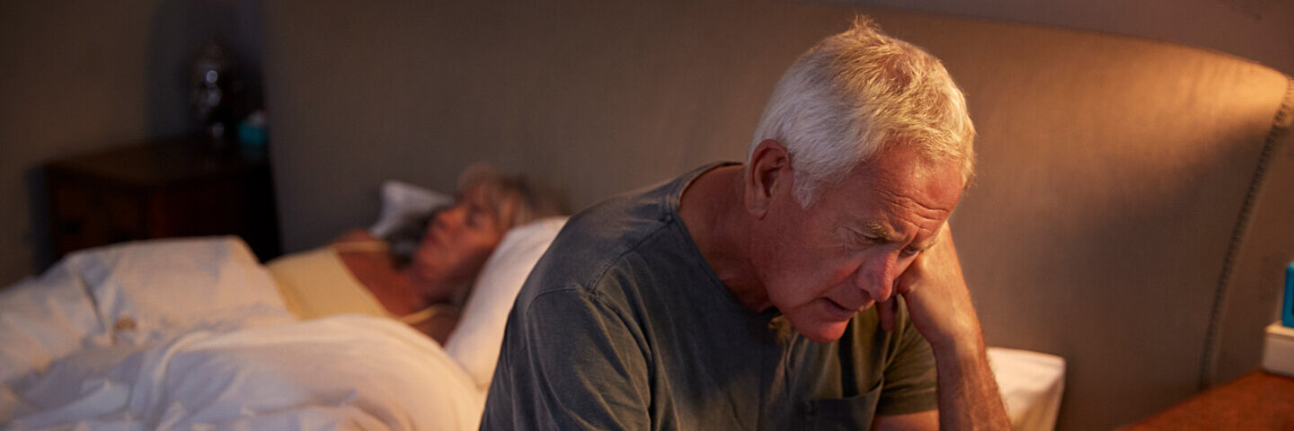 Ein älterer Mann sitzt mit leicht gesenktem Kopf auf einem Bett und stützt den linken Arm ab.