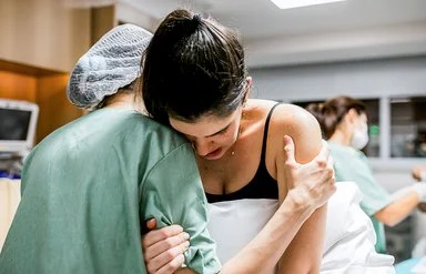 Eine Gebärende lehnt ihren Kopf an die Schulter einer Hebamme und hält sich dabei mit einer Hand an ihrem rechten Oberarm fest, während die Hebamme die Schwangere an deren linkem Oberarm stützt.