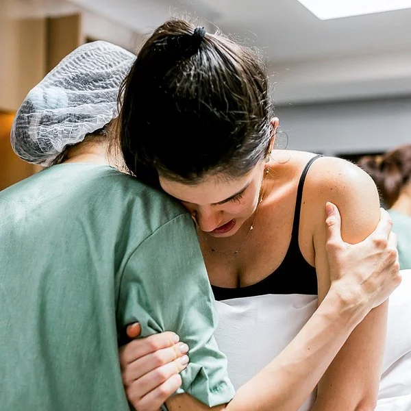 Eine Gebärende lehnt ihren Kopf an die Schulter einer Hebamme und hält sich dabei mit einer Hand an ihrem rechten Oberarm fest, während die Hebamme die Schwangere an deren linkem Oberarm stützt.