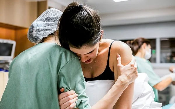 Eine Gebärende lehnt ihren Kopf an die Schulter einer Hebamme und hält sich dabei mit einer Hand an ihrem rechten Oberarm fest, während die Hebamme die Schwangere an deren linkem Oberarm stützt.