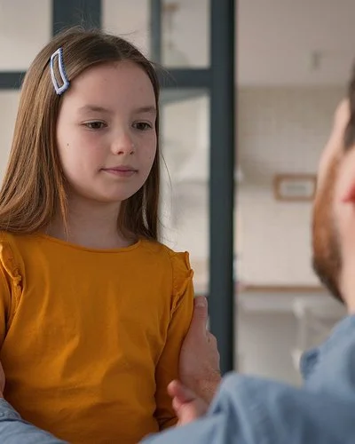 Ein Mädchen steht einem Mann gegenüber. Der Mann ist nur von hinten zu sehen. Er kniet und fasst das Kind an die Schultern. Das Mädchen blickt nach unten. Sein Mund ist geschlossen.