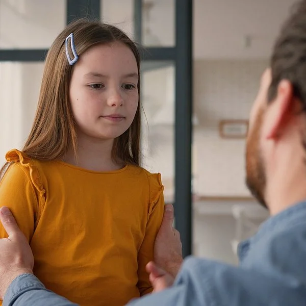 Ein Mädchen steht einem Mann gegenüber. Der Mann ist nur von hinten zu sehen. Er kniet und fasst das Kind an die Schultern. Das Mädchen blickt nach unten. Sein Mund ist geschlossen.