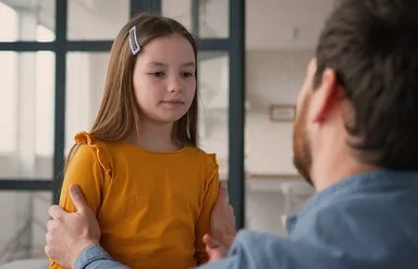 Ein Mädchen steht einem Mann gegenüber. Der Mann ist nur von hinten zu sehen. Er kniet und fasst das Kind an die Schultern. Das Mädchen blickt nach unten. Sein Mund ist geschlossen.