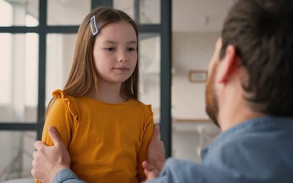 Ein Mädchen steht einem Mann gegenüber. Der Mann ist nur von hinten zu sehen. Er kniet und fasst das Kind an die Schultern. Das Mädchen blickt nach unten. Sein Mund ist geschlossen.