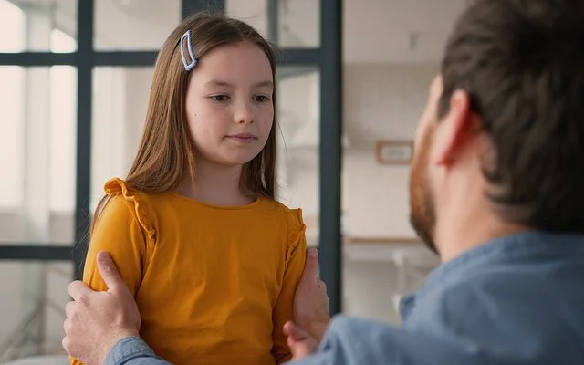 Ein Mädchen steht einem Mann gegenüber. Der Mann ist nur von hinten zu sehen. Er kniet und fasst das Kind an die Schultern. Das Mädchen blickt nach unten. Sein Mund ist geschlossen.