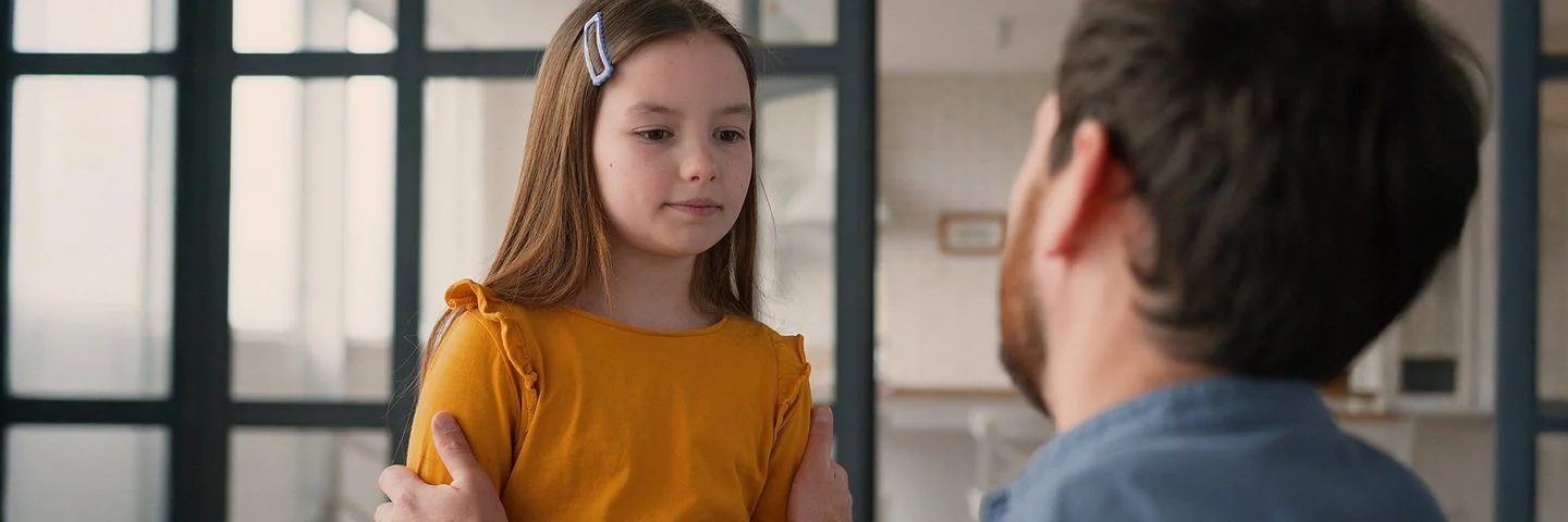 Ein Mädchen steht einem Mann gegenüber. Der Mann ist nur von hinten zu sehen. Er kniet und fasst das Kind an die Schultern. Das Mädchen blickt nach unten. Sein Mund ist geschlossen.
