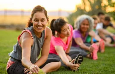 Mehrere Erwachsene dehnen sich nach dem Training auf einer Wiese; Outdoor-Sport im Sommer bei warmem Abendlicht.