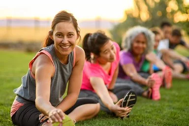 Mehrere Erwachsene dehnen sich nach dem Training auf einer Wiese; Outdoor-Sport im Sommer bei warmem Abendlicht.