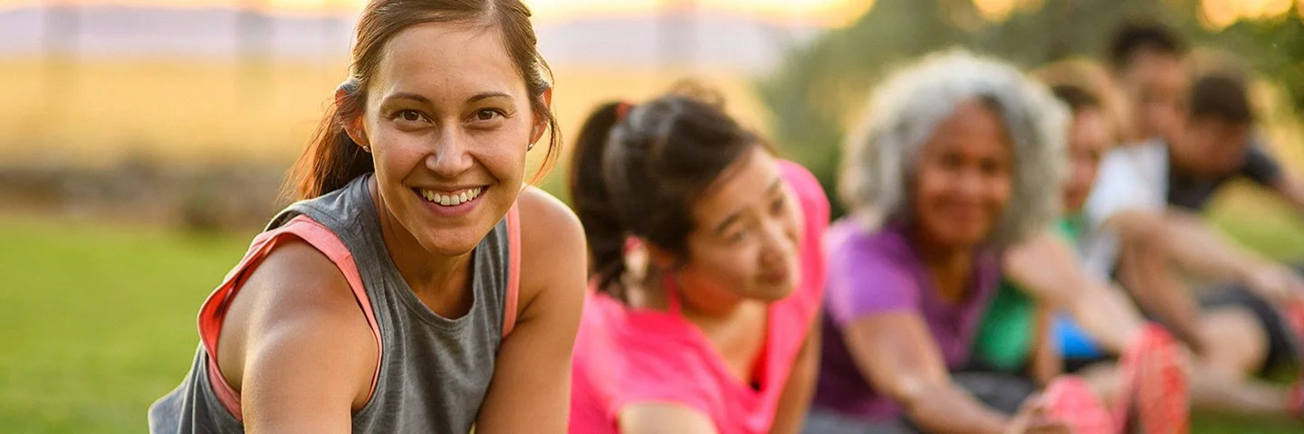 Mehrere Erwachsene dehnen sich nach dem Training auf einer Wiese; Outdoor-Sport im Sommer bei warmem Abendlicht.