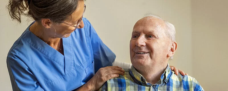 Eine Pflegerin spricht mit einem pflegebedürftigen alten Mann. Dabei berührt sie ihn mit beiden Händen an seinen Schultern.