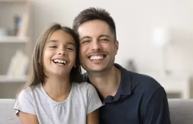 Ein Vater und seine Tochter lachen in die Kamera und zeigen dabei die Zähne.