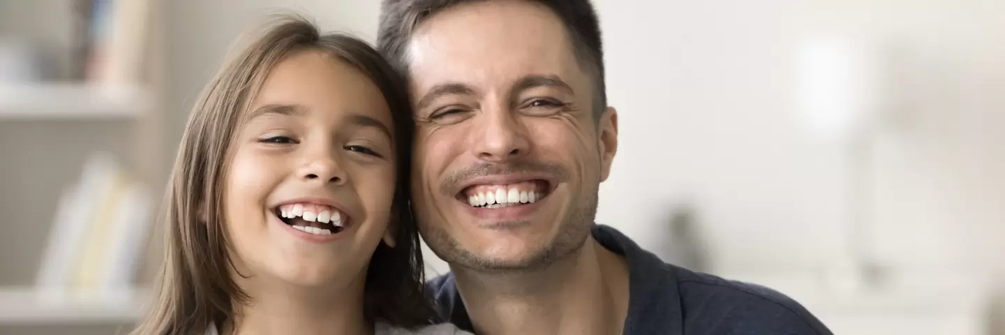 Ein Vater und seine Tochter lachen in die Kamera und zeigen dabei die Zähne.