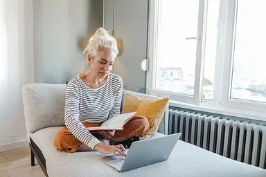 Eine Frau mit hochgesteckten Haaren sitzt im Schneidersitz auf einem grauen Sofa. Sie trägt ein schwarz-weiß gestreiftes Langarmshirt und eine orangene Hose. Mit einer Hand tippt sie auf dem Laptop vor sich, mit der anderen hält sie ein Buch.