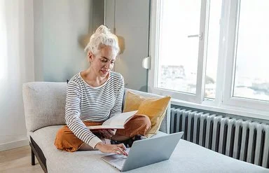 Eine Frau mit hochgesteckten Haaren sitzt im Schneidersitz auf einem grauen Sofa. Sie trägt ein schwarz-weiß gestreiftes Langarmshirt und eine orangene Hose. Mit einer Hand tippt sie auf dem Laptop vor sich, mit der anderen hält sie ein Buch.