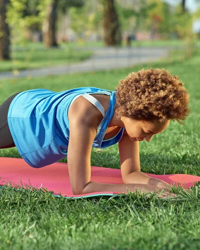 Eine junge Frau in Sportbekleidung macht sogenannte Planks auf einer Yogamatte in einem Park.