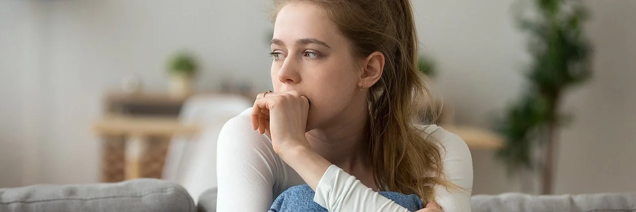 Eine junge Frau sitzt mit angezogenen Beinen auf der Couch und guckt nachdenklich.