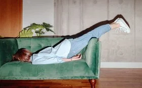 Frau liegt mit dem Oberkörper auf einem grünen Sofa und streckt die Beine in Jeans und weißen Sneakern nach oben aus.