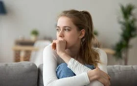 Eine junge Frau sitzt mit angezogenen Beinen auf der Couch und guckt nachdenklich.