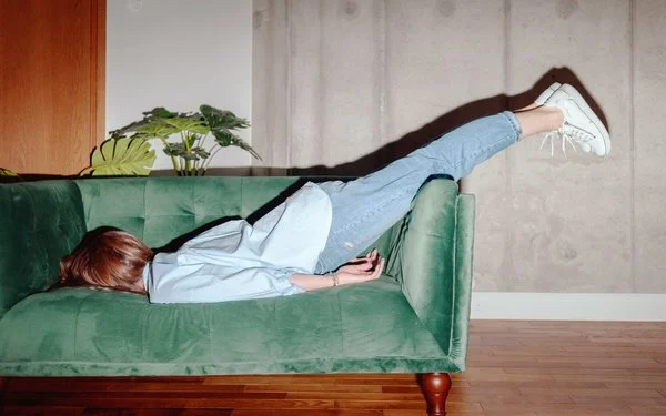 Frau liegt mit dem Oberkörper auf einem grünen Sofa und streckt die Beine in Jeans und weißen Sneakern nach oben aus.