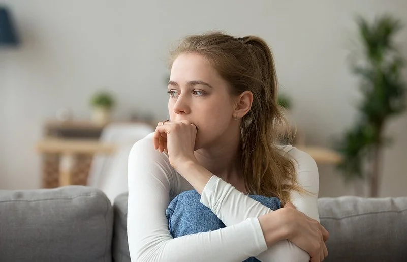 Eine junge Frau sitzt mit angezogenen Beinen auf der Couch und guckt nachdenklich.