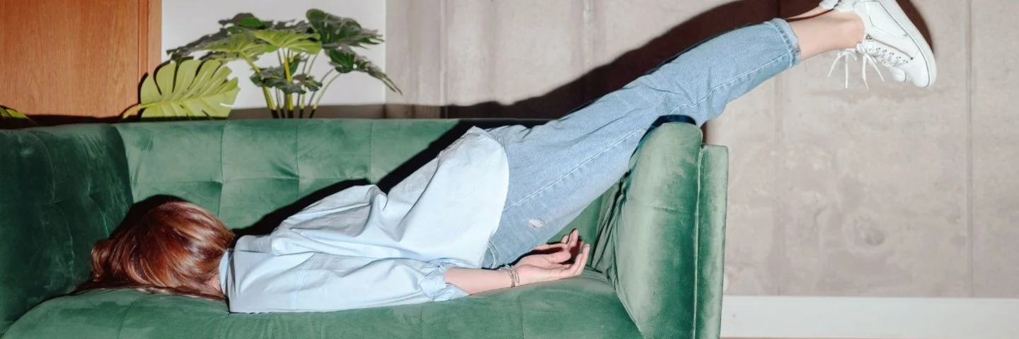 Frau liegt mit dem Oberkörper auf einem grünen Sofa und streckt die Beine in Jeans und weißen Sneakern nach oben aus.