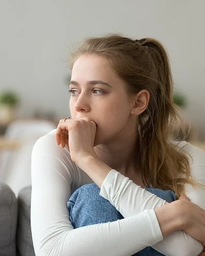 Eine junge Frau sitzt mit angezogenen Beinen auf der Couch und guckt nachdenklich.