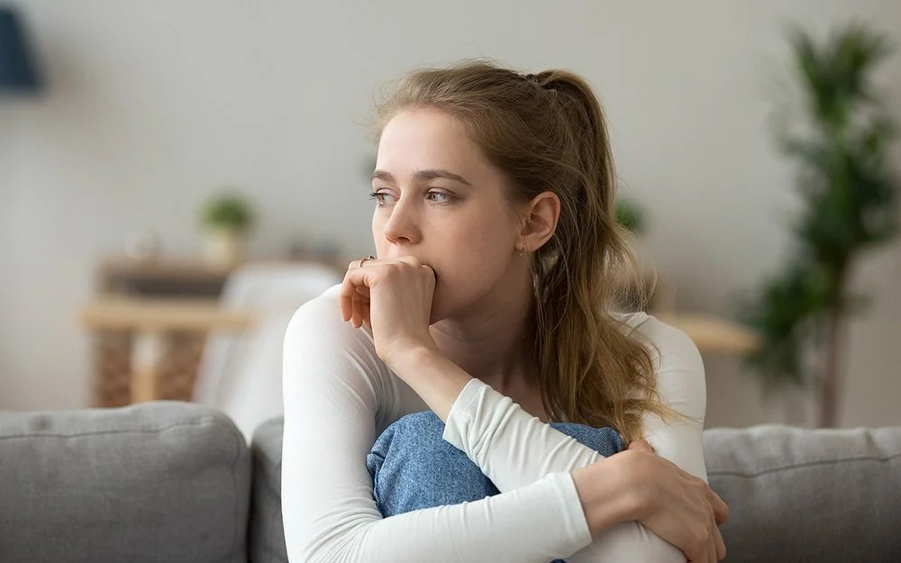 Eine junge Frau sitzt mit angezogenen Beinen auf der Couch und guckt nachdenklich.