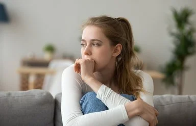 Eine junge Frau sitzt mit angezogenen Beinen auf der Couch und guckt nachdenklich.