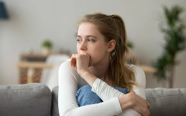 Eine junge Frau sitzt mit angezogenen Beinen auf der Couch und guckt nachdenklich.
