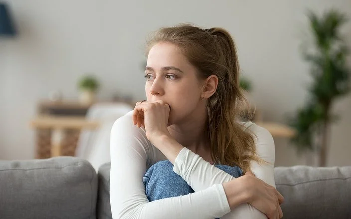 Eine junge Frau sitzt mit angezogenen Beinen auf der Couch und guckt nachdenklich.