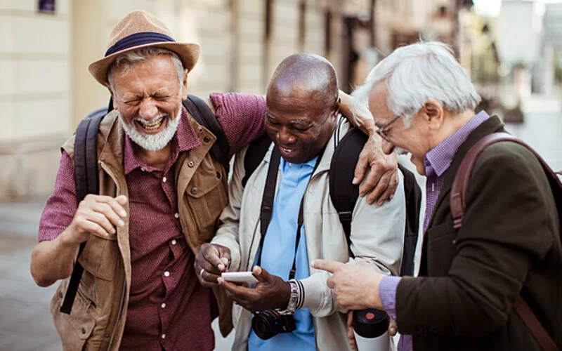 Ein älterer Herr zeigt seinen Freunden etwas auf dem Smartphone, worüber alle lachen. Digitalisierung im Alter kann Spaß machen.
