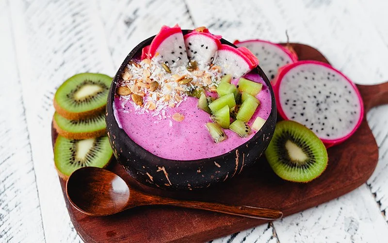 Eine Schüssel mit einem Bowl-Gericht steht auf einem Holzbrett, daneben liegen ein Holzlöffel, eine aufgeschnittene Kiwi und eine Drachenfrucht.