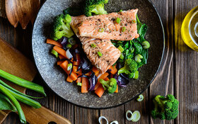 Gesunder Fisch (Lachs) mit gedünsteten Karotten, Brokkoli, Zwiebeln und Spinat auf einem Teller.