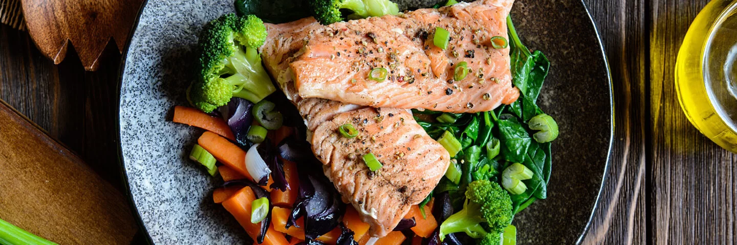 Gesunder Fisch (Lachs) mit gedünsteten Karotten, Brokkoli, Zwiebeln und Spinat auf einem Teller.