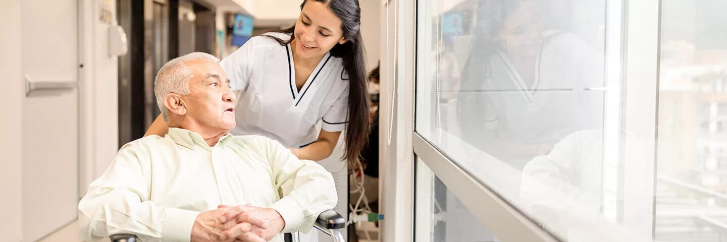 3)	Eine weibliche Pflegefachperson fährt einen Patienten im Rollstuhl durch den Flur.