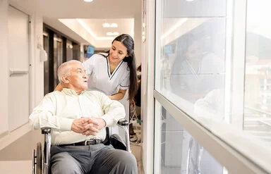 3)	Eine weibliche Pflegefachperson fährt einen Patienten im Rollstuhl durch den Flur.