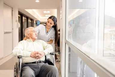 3)	Eine weibliche Pflegefachperson fährt einen Patienten im Rollstuhl durch den Flur.