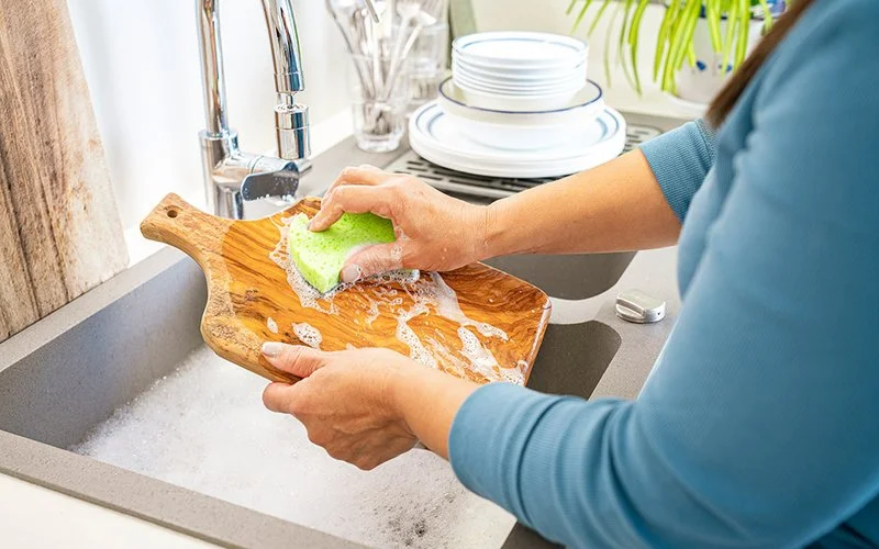 Hände, die ein Schneidebrett mit einem Schwamm über einem mit Wasser gefüllten Spülbecken reinigen.