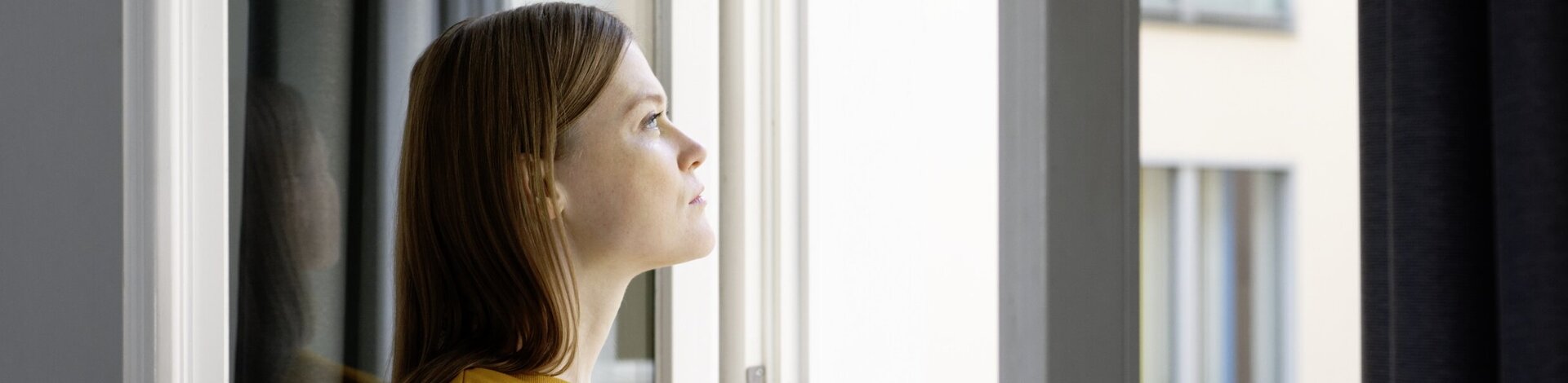 Frau mit gelbem Pulli guckt nachdenklich und schweremutig aus dem Fenster.