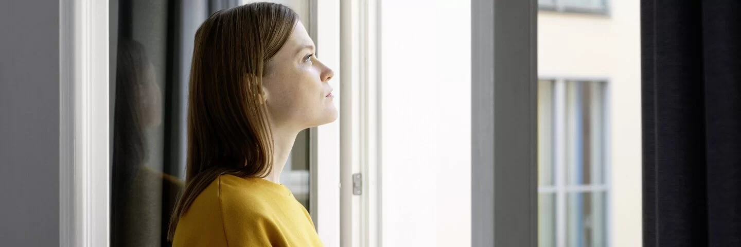 Frau mit gelbem Pulli guckt nachdenklich und schweremutig aus dem Fenster.