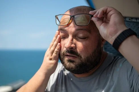 Ein Mann mit Bart nimmt bei sonnigem Wetter seine Brille ab. Er hat die Augen geschlossen und die Hände an den Schläfen.