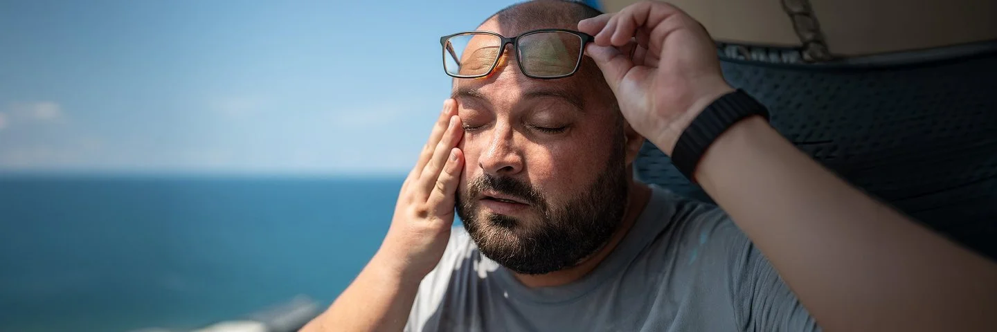 Ein Mann mit Bart nimmt bei sonnigem Wetter seine Brille ab. Er hat die Augen geschlossen und die Hände an den Schläfen.