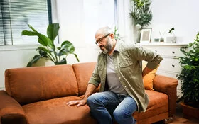 Ein Mann in mittlerem Alter sitzt auf dem Sofa und hält eine Hand ins Kreuz.