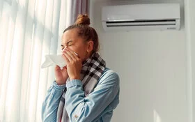 Niesende Frau, die ein Taschentuch vor ihr Gesicht hält, in einem hellen Raum mit zugezogenen Vorhängen und Klimaanlage an der Wand.