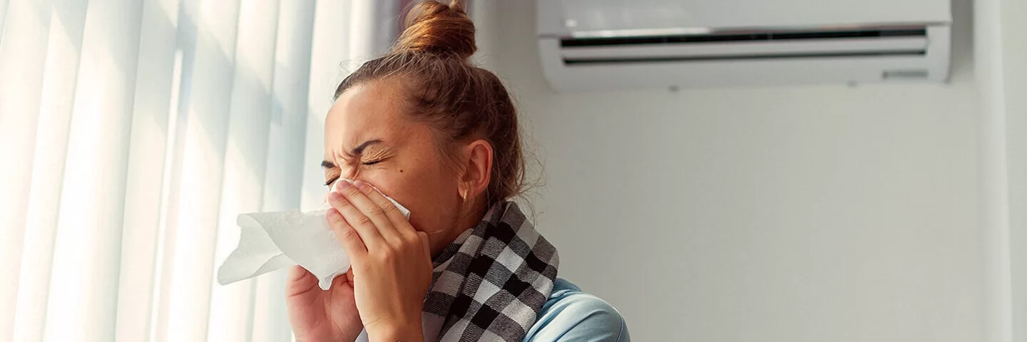 Niesende Frau, die ein Taschentuch vor ihr Gesicht hält, in einem hellen Raum mit zugezogenen Vorhängen und Klimaanlage an der Wand.