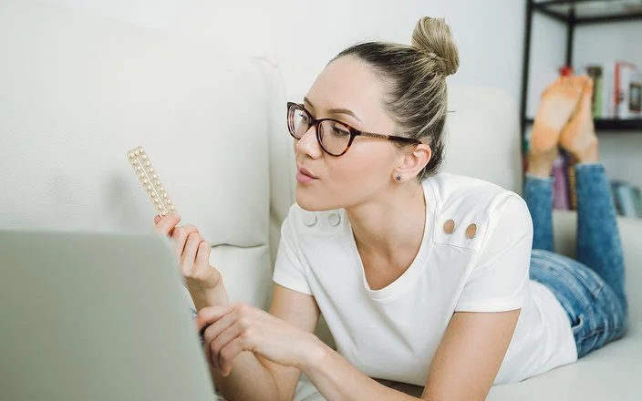 Junge Frau mit Brille liegt auf dem Bett und betrachtet nachdenklich eine Blisterpackung mit Tabletten, während ein Laptop vor ihr steht.