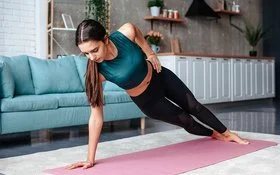 Eine Frau in einem dunkelgrünen Oberteil führt in ihrem Wohnzimmer eine Seitenplanke aus.