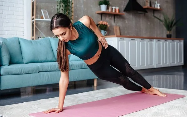 Eine Frau in einem dunkelgrünen Oberteil führt in ihrem Wohnzimmer eine Seitenplanke aus.